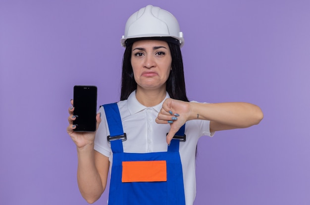 Young builder woman in construction uniform and safety helmet showing smartphone looking at front being displeased showing thumb down standing over purple wall