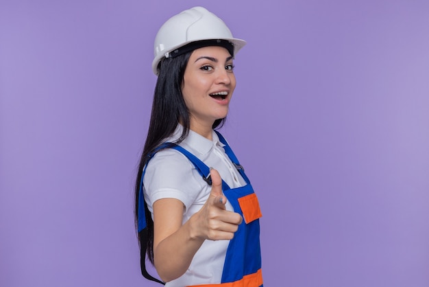 Giovane donna del costruttore in uniforme da costruzione e casco di sicurezza che sorride allegramente indicando con il dito indice davanti in piedi sopra la parete viola