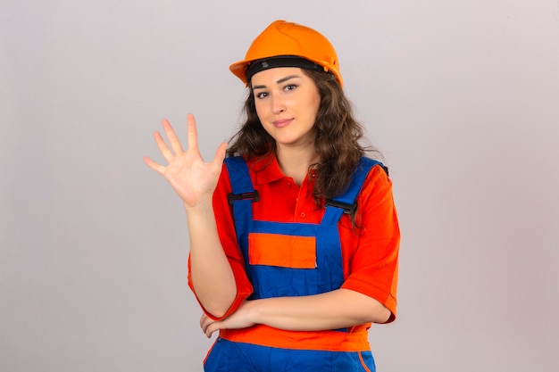 Young builder woman in construction uniform and safety helmet smiling cheerful showing and pointing up with fingers number five looking confident and happy over isolated white wall