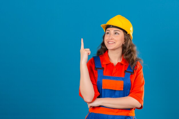 Young builder woman in construction uniform and safety helmet smiling cheerfully looking confident pointing up with index finger over isolated blue wall