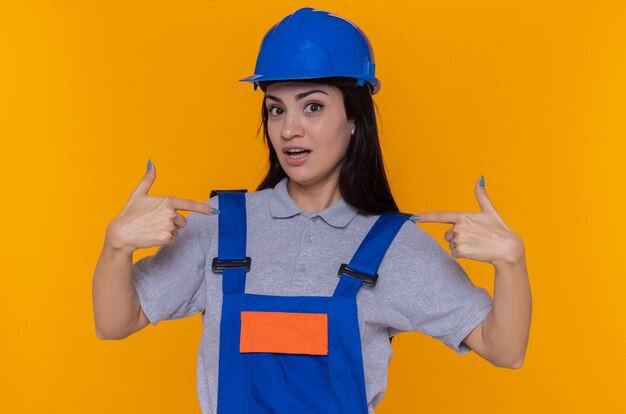 オレンジ色の壁の上に立っている自分自身を指して幸せそうに見えて驚いて笑顔の建設制服と安全ヘルメットの若いビルダーの女性