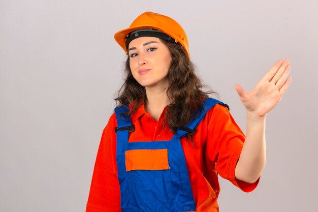 Young builder woman in construction uniform and safety helmet smiling waving hand welcoming and greeting gesture over isolated white wall