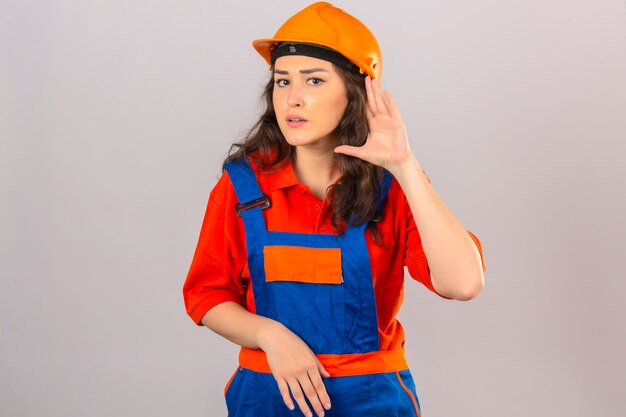 Young builder woman in construction uniform and safety helmet smiling with hand over ear listening an hearing to rumor or gossip over isolated white wall