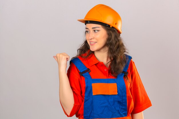 Young builder woman in construction uniform and safety helmet smiling with happy face looking and pointing to the side with thumb up over isolated white wall