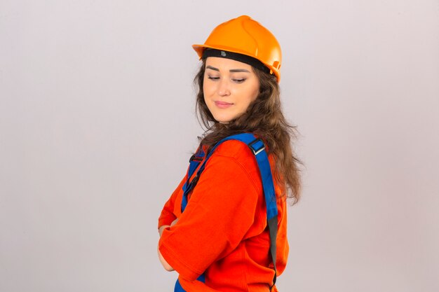 Young builder woman in construction uniform and safety helmet standing sideways looking down over isolated white wall