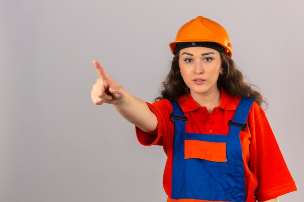 Young builder woman in construction uniform and safety helmet standing with finger up warning of danger over isolated white wall