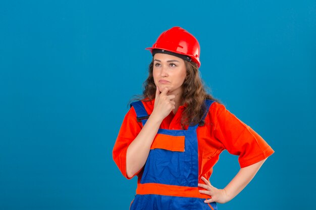 Young builder woman in construction uniform and safety helmet standing with hand on chin thinking having doubts over isolated blue wall