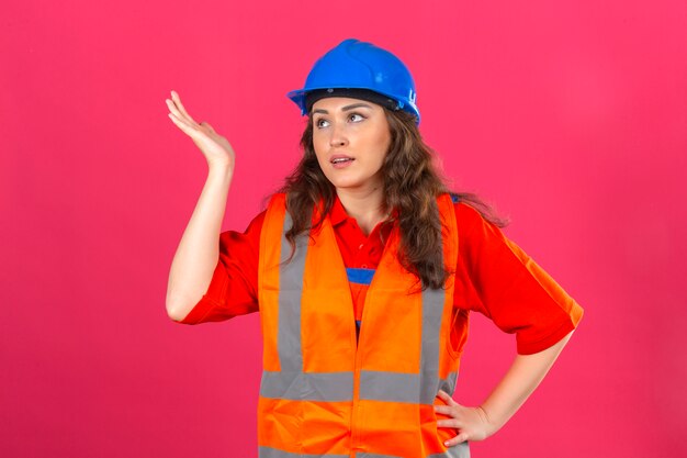 Young builder woman in construction uniform and safety helmet standing with hand raised gesturing disappointed having no idea concept over isolated pink wall