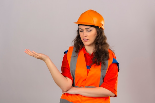 Young builder woman in construction uniform and safety helmet standing with hand raised thinking trying to make choice having doubts over isolated white wall