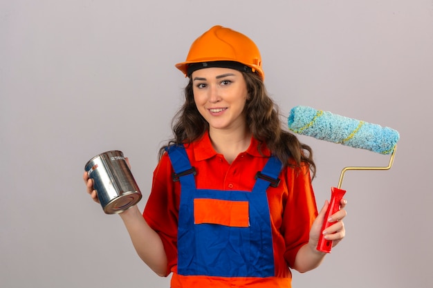 Young builder woman in construction uniform and safety helmet standing with paint roller and paint can smiling cheerfully over isolated white wall