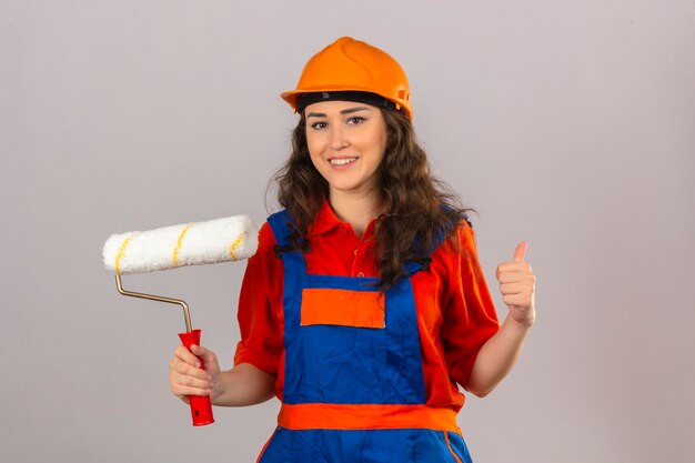 Young builder woman in construction uniform and safety helmet standing with paint roller smiling showing thumbs up over isolated white wall