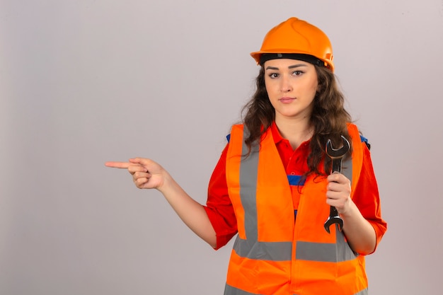 Free photo young builder woman in construction uniform and safety helmet standing with wrench pointing index finger to the side over isolated white wall