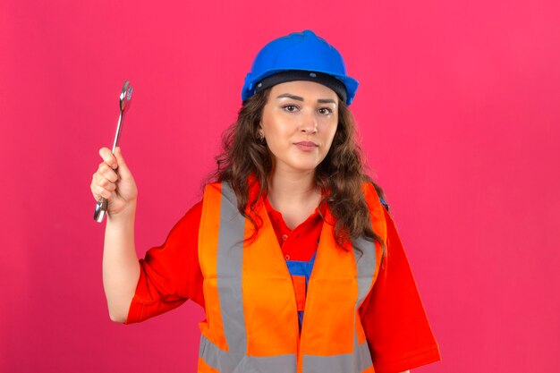 Молодой строитель женщина в строительной форме и защитный шлем, стоя с гаечным ключом, улыбаясь на изолированных розовые стены