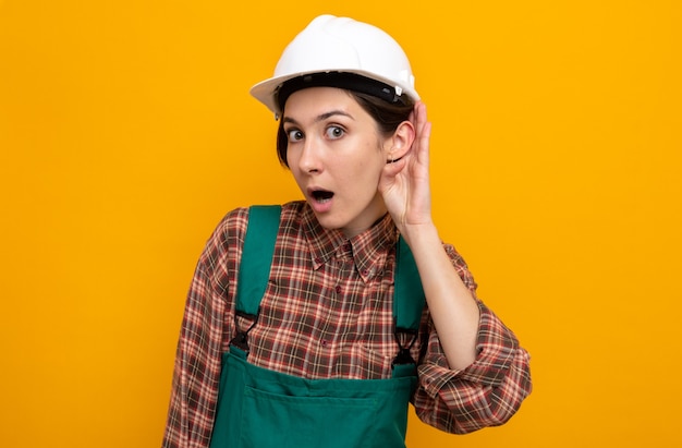 Young builder woman in construction uniform and safety helmet surprised holding hand on ear trying to listen to gossip standing on orange