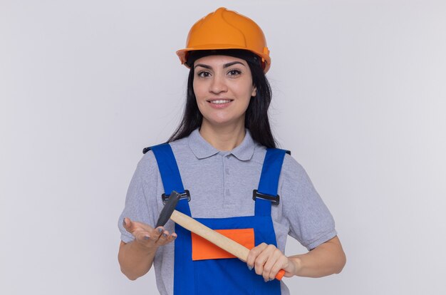 Young builder woman in construction uniform and safety helmet swinging a hammer looking at front smiling confident standing over white wall