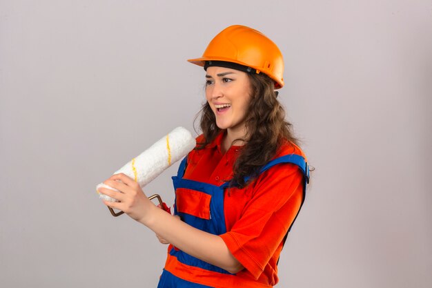 Young builder woman in construction uniform and safety helmet using paint roller as microphone singing song fun at work concept over isolated white wall