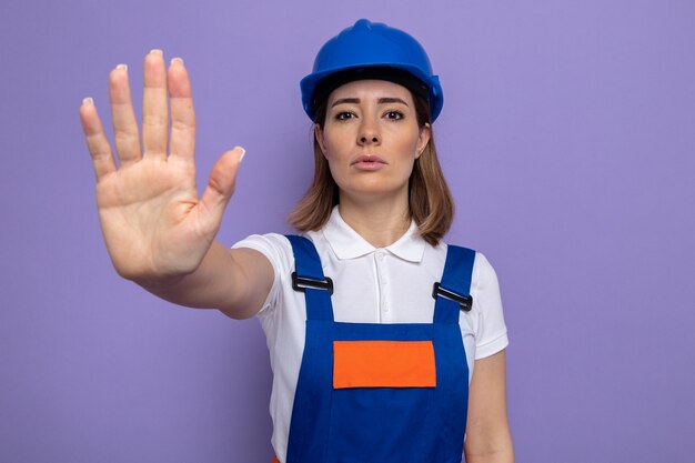 Young builder woman in construction uniform and safety helmet  with serious face making stop gesture with hand standing over purple wall