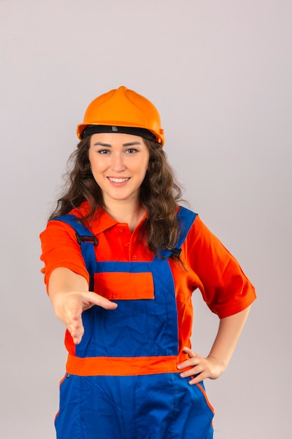 Молодая женщина строитель в строительной форме и защитный шлем с улыбкой, предлагая руку, делая приветствие жест над изолированной белой стене