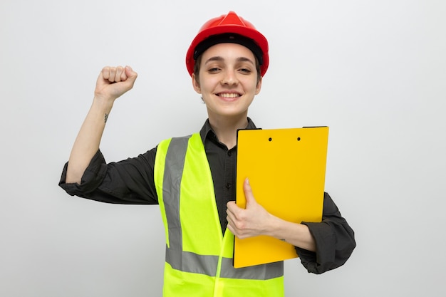 Молодая женщина-строитель в строительном жилете и защитном шлеме держит буфер обмена счастливым и взволнованным сжимающим кулаком, стоящим над белой стеной