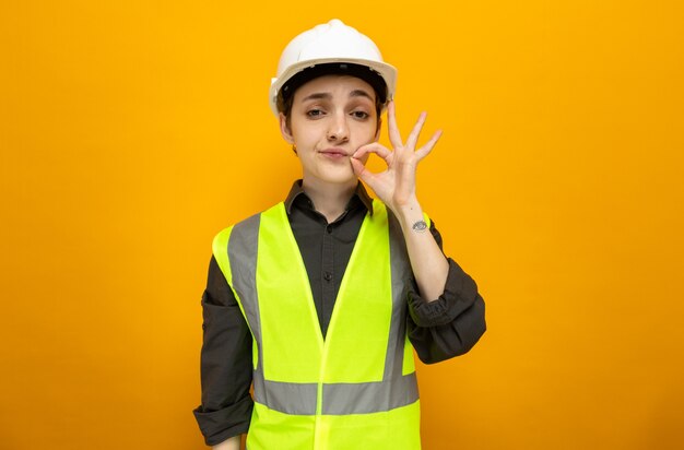 オレンジ色の上に立っているジッパーで口を閉じるような沈黙のジェスチャーを作る建設ベストと安全ヘルメットの若いビルダーの女性