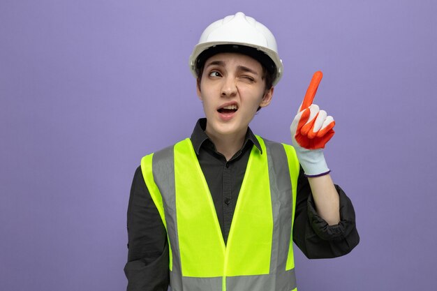 Молодая женщина-строитель в строительном жилете и защитном шлеме в резиновых перчатках смущенно смотрит вверх, показывая сомневающийся указательный палец, стоящий над фиолетовой стеной