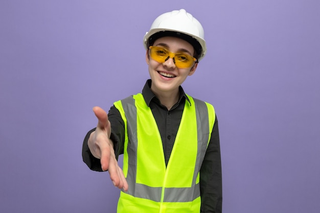 Молодой строитель женщина в строительном жилете и защитном шлеме в защитных желтых очках улыбается дружелюбно, предлагая рукой приветствующий жест, стоящий над синей стеной
