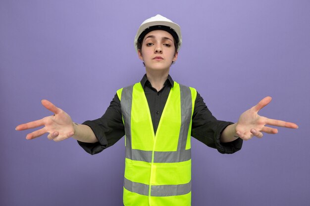 Молодая женщина-строитель в строительном жилете и защитном шлеме с серьезным лицом, недовольно поднимающим руки, стоя на фиолетовом