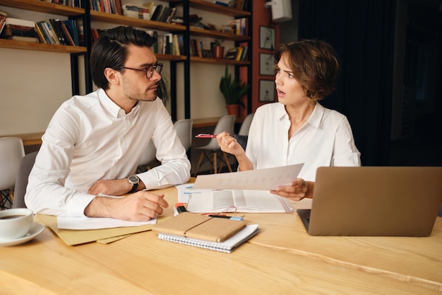 Foto gratuita giovani colleghi di lavoro che discutono emotivamente di un nuovo progetto seduti al tavolo con laptop e documenti in un ufficio moderno