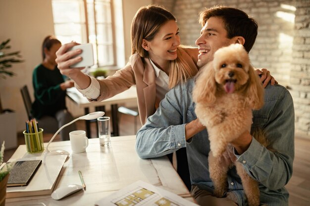 オフィスで犬と一緒に自分撮りをしながら楽しんでいる若いビジネスカップル