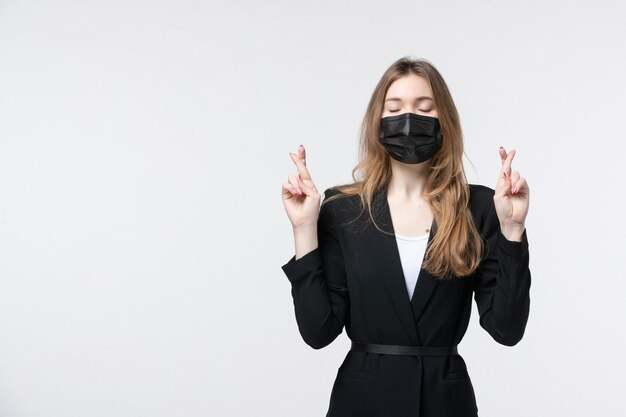 Young business lady in suit wearing surgical mask and crossing her fingers by closing her eyes on isolated white wall