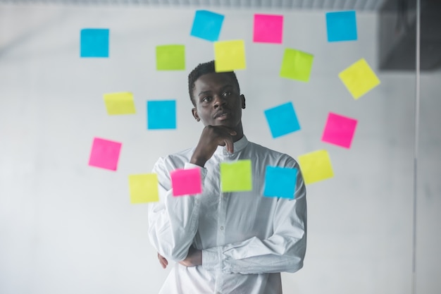 Free photo young business man standing in front of stickers glass wall and looking on futures plans at his office place