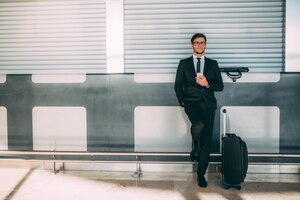 Бесплатное фото Молодой бизнесмен стоит у телефона с чемоданом в аэропорту в ожидании рейса xaxa