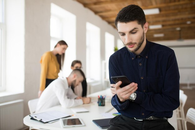 バックグラウンドで同僚とオフィスで思慮深く携帯電話を使用して暗いシャツのひげを持つ若いビジネスマン