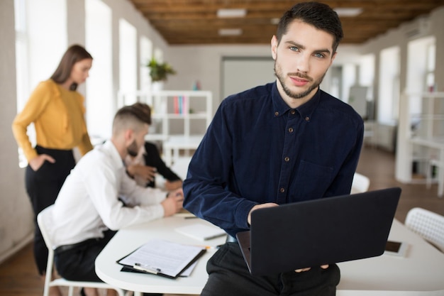 Молодой деловой человек с бородой в рубашке задумчиво смотрит в камеру, держа ноутбук в руках, проводя время в офисе с коллегами на заднем плане
