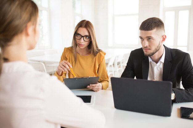 Young business man with laptop and business woman in eyeglasses thoughtfully looking at applicant Young employers spending job interview in modern office