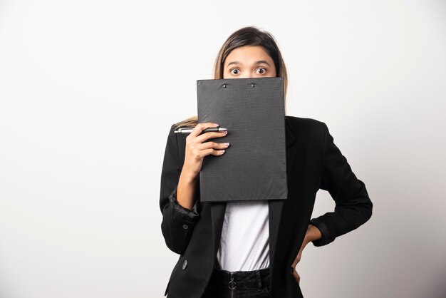 Young business woman covering her face with clipboard on white wall