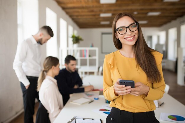 Young business woman in eyeglasses happily looking in camera while holding cellphone in hands in office with colleagues on background