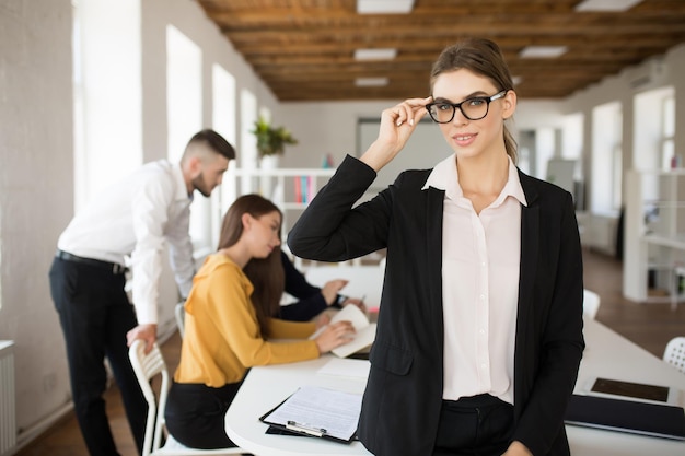 Foto gratuita giovane donna d'affari in occhiali e camicia che guarda sognante a porte chiuse mentre trascorre del tempo in ufficio con i colleghi sullo sfondo