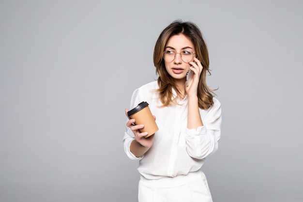 若いビジネス女性が灰色の壁に分離されたコーヒーを飲みながら携帯電話で話しているポーズ