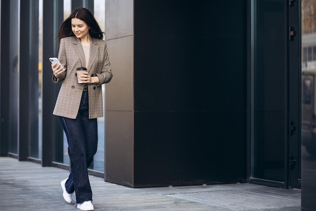 Young business woman walking by the office center using phone and drinking coffee