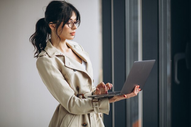 オフィスの窓のそばに立っているラップトップを持つ若いビジネス女性