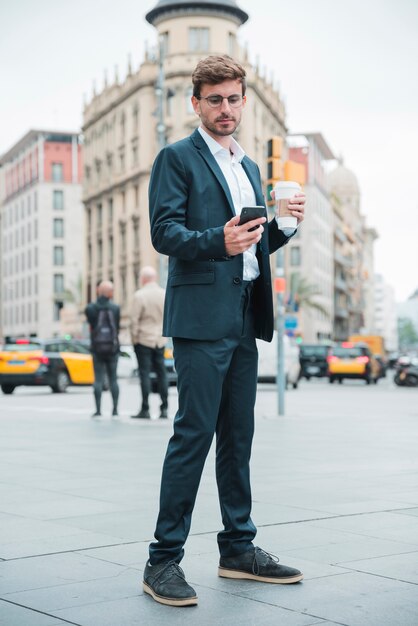 Young businessman holding coffee cup standing on street looking at mobile phone