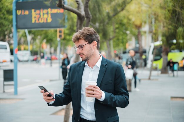 Молодой бизнесмен держит одноразовые чашки кофе, глядя на мобильный телефон