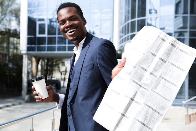 Free photo young businessman holding disposable coffee cup showing newspaper toward camera