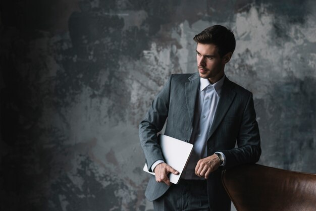 Young businessman leaning on chair holding laptop in his hand against grunge background