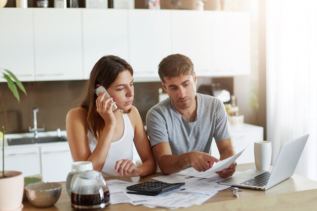 Young businessman pointing with finger at document, trying to expain something to his wife who is speaking over smart phone. Couple reviewing their bank accounts and calculating annual figures