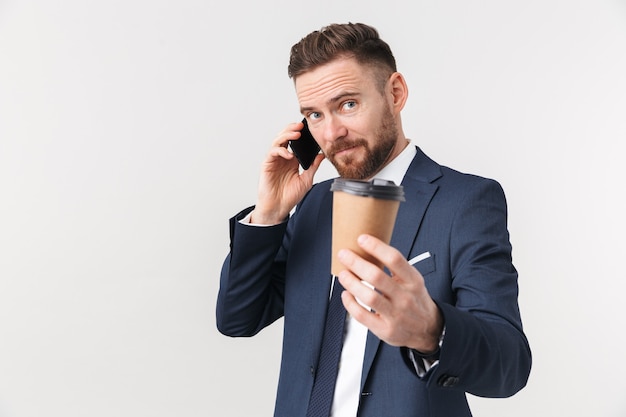 Молодой бизнесмен позирует изолированно над белой стеной, пить кофе, разговаривает по телефону