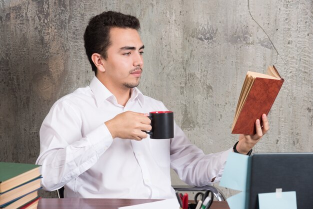 Молодой бизнесмен читает книгу и пьет чай за столом.