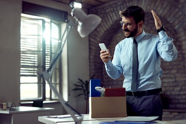 Young businessman reading text message on cell phone while feeling angry about losing his job in the office