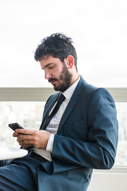Молодой бизнесмен, сидя в офисе с помощью мобильного телефона